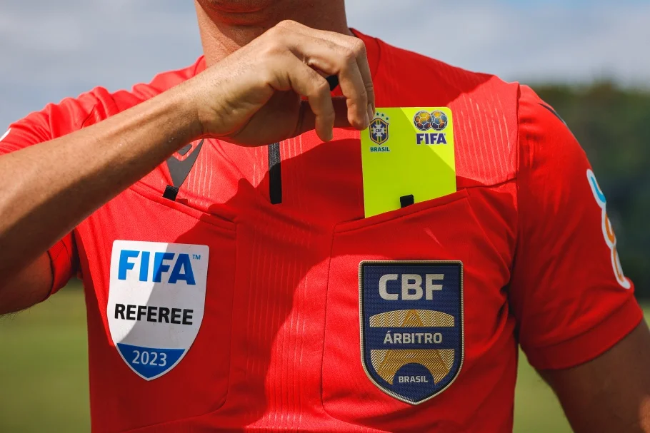 Fotografia em close do tronco de um árbitro de futebol brasileiro vestido com sua camisa de uniforme vermelha oficial (Futebol-Arbitragem-FOTO-Rodrigo-Ferreira-CBF.webp). O árbitro, cujo rosto não é visível, tem a mão direita levantada, retirando o cartão amarelo do bolso do peito. O uniforme exibe com destaque os emblemas da FIFA (árbitro de 2023), da CBF e da Associação Brasileira de Árbitros de Futebol (ABAF). O fundo desfocado sugere um campo de futebol em um dia de sol. Essa imagem é comumente usada para ilustrar artigos e discussões sobre o mundo da arbitragem, incluindo tópicos como quanto ganha um árbitro no futebol brasileiro.
